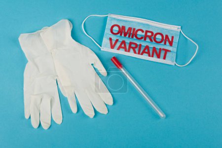 Top view of medical mask with omicron variant near cotton swab and latex gloves on blue background 
