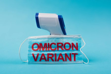 Medical mask with omicron variant lettering near pyrometer on blue background 