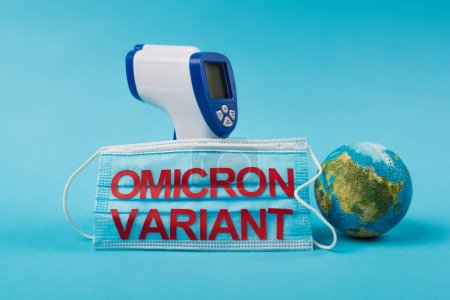 Pyrometer near medical mask with omicron variant lettering and globe on blue background 