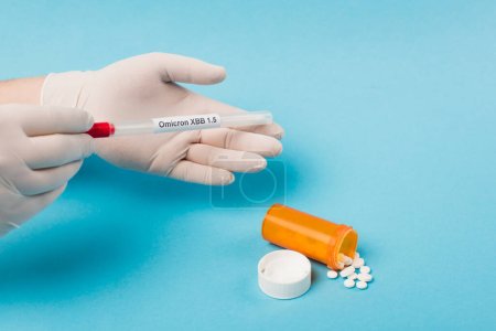 Cropped view of doctor in latex gloves holding cotton swab with omicron lettering near pills on blue background 