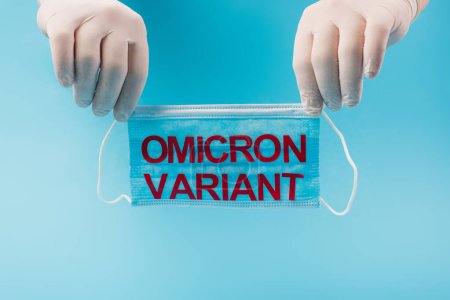 Cropped view of doctor in latex gloves holding medical mask with omicron variant lettering isolated on blue 