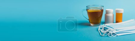 Medical masks near pills and cup of tea with lemon on blue background, banner 