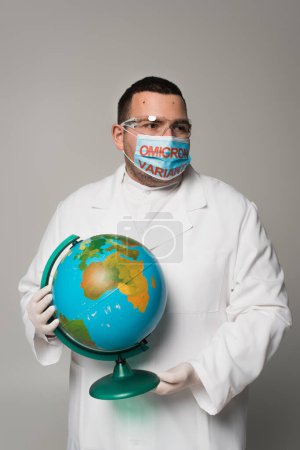 Scientist in medical mask with omicron variant lettering holding globe isolated on grey 