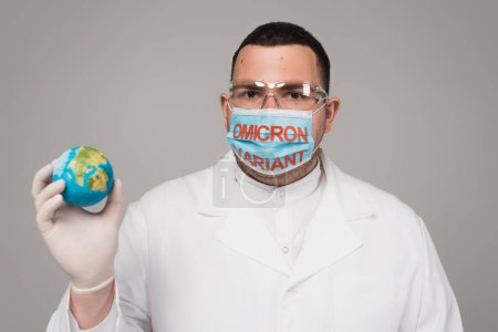 Scientist in medical mask with omicron variant holding small globe isolated on grey 