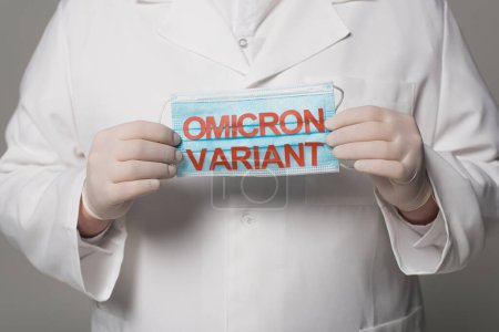 Cropped view of doctor in latex gloves holding medical mask with omicron variant lettering on grey background 