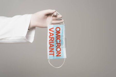 Cropped view of scientist in latex glove holding medical mask with omicron variant lettering isolated on grey 