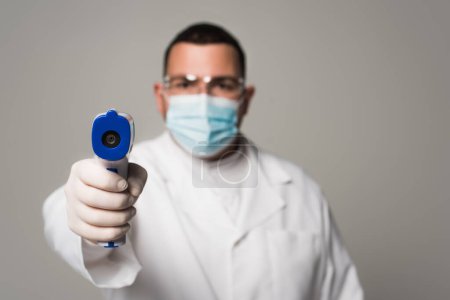 Téléchargez les photos : Médecin flou dans le masque médical tenant pyromètre isolé sur gris - en image libre de droit