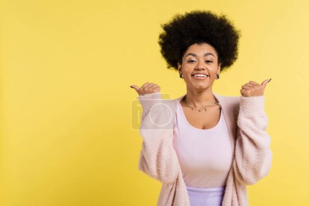 Téléchargez les photos : Smiling african american woman in cardigan pointing with thumbs and looking at camera isolated on yellow - en image libre de droit