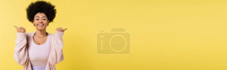 Photo for African american woman in cardigan and top pointing with thumbs and smiling at camera isolated on yellow, banner - Royalty Free Image