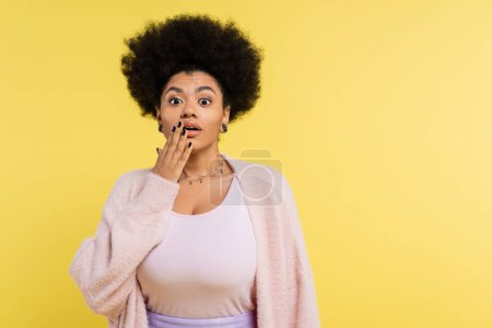Photo for Astonished african american woman in top and soft cardigan covering mouth with hand isolated on yellow - Royalty Free Image