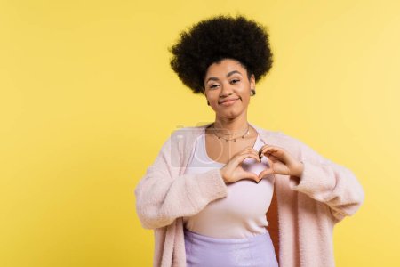 Foto de Positive african american woman in soft cardigan showing heart sign and looking at camera isolated on yellow - Imagen libre de derechos