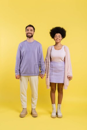 full length of trendy interracial couple holding hands and smiling at camera on yellow background