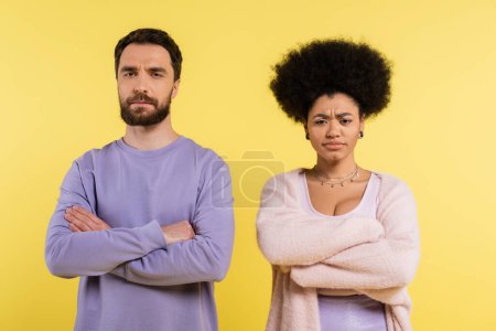 Foto de Displeased interracial couple standing with crossed arms while looking at camera isolated on yellow - Imagen libre de derechos