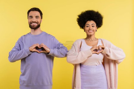 happy multiethnic couple looking at camera and showing heart signs isolated on yellow