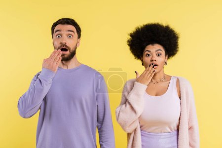 thrilled interracial couple covering open mouth with hands and looking at camera isolated on yellow