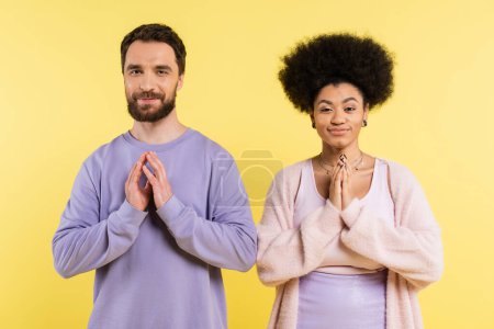 cunning interracial couple smiling at camera and showing please gesture isolated on yellow