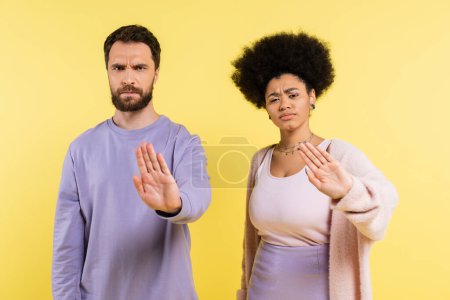 displeased and frowning multiethnic couple looking at camera and showing refuse gesture isolated on yellow