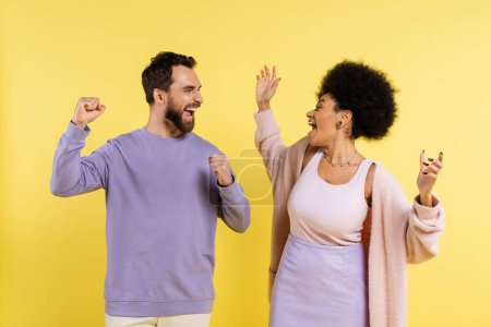 overjoyed multiethnic couple screaming and showing win gesture while looking at each other isolated on yellow