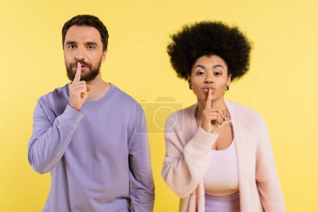 stylish multiethnic couple looking at camera and showing hush sign isolated on yellow