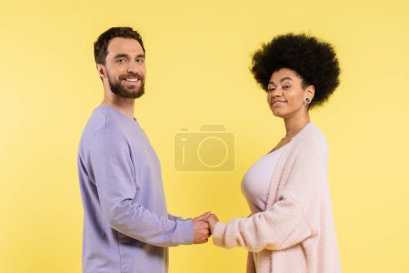 cheerful multiethnic couple holding hands and looking at camera while posing isolated on yellow