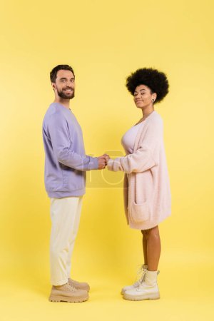 Foto de Full length of interracial couple in stylish clothes holding hands and looking at camera on yellow background - Imagen libre de derechos