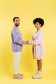 full length of interracial couple in stylish clothes holding hands and looking at camera on yellow background mug #635603980
