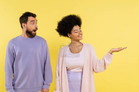 smiling african american woman pointing with hand near amazed bearded man isolated on yellow