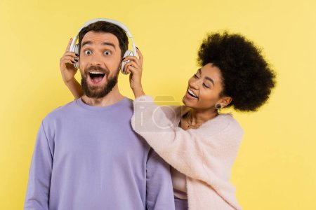 cheerful african american woman putting wireless headphones on excited bearded man isolated on yellow