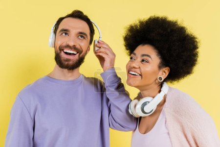 smiling african american woman looking at happy man in wireless headphones isolated on yellow