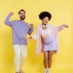 full length of cheerful interracial couple in trendy clothes and wireless headphones dancing on yellow 