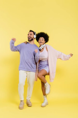full length of excited and stylish multiethnic couple dancing in wireless headphones on yellow background