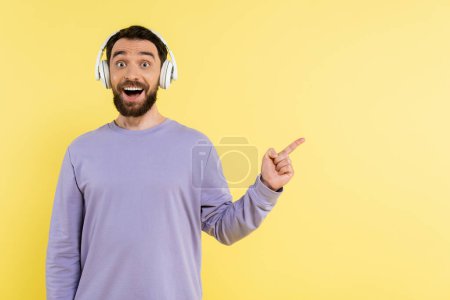 amazed man in wireless headphones pointing with finger and looking at camera isolated on yellow