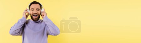 Foto de Cheerful man with closed eyes adjusting wireless headphones and laughing isolated on yellow, banner - Imagen libre de derechos