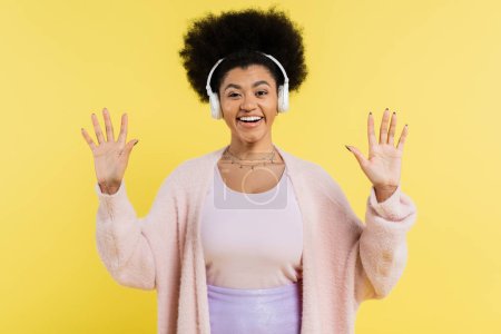 Photo for Optimistic african american woman in wireless headphones and cardigan waving hands and looking at camera isolated on yellow - Royalty Free Image