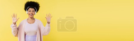 joyful african american woman listening music in wireless headphones and waving hands isolated on yellow, banner