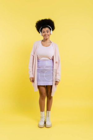 full length of cheerful african american woman in cardigan and wireless headphones looking at camera on yellow background