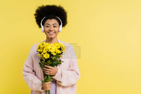 pleased african american woman in wireless headphones smiling at camera while holding flowers isolated on yellow