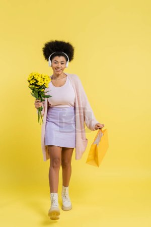 full length of smiling african american woman in wireless headphones walking with flowers and shopping bag on yellow background