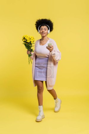 full length of happy african american woman in wireless headphones walking with bouquet and smartphone on yellow 