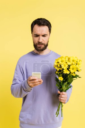 thoughtful and frowning man with flowers using mobile phone isolated on yellow