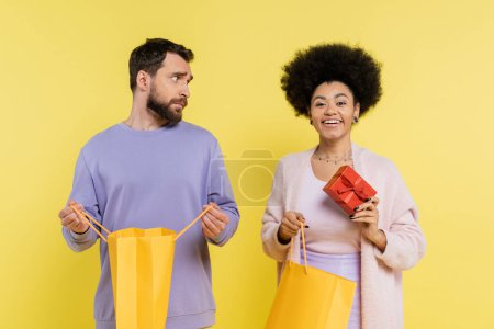 happy african american woman holding gift box near offended man with shopping bag isolated on yellow