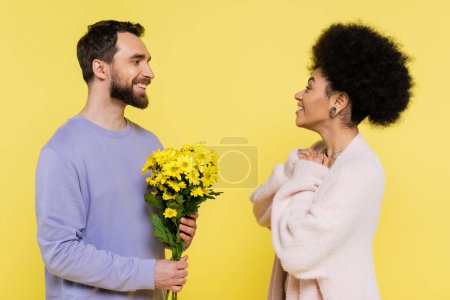happy bearded man holding bouquet near amazed african american woman isolated on yellow