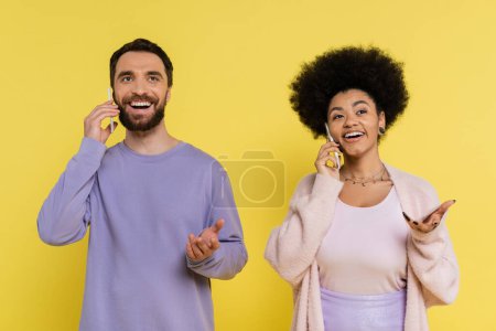 trendy and positive interracial couple gesturing while talking on mobile phones isolated on yellow