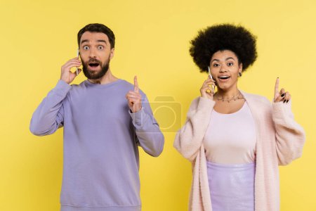 Photo for Amazed multiethnic couple showing idea gesture while talking on smartphones isolated on yellow - Royalty Free Image