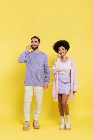 full length of smiling interracial couple in stylish clothes talking on smartphones on yellow background