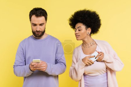 curious african american woman looking at boyfriend messaging on mobile phone isolated on yellow
