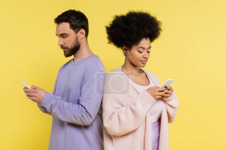 trendy interracial couple messaging on mobile phones while standing back to back isolated on yellow