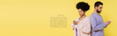 stylish interracial couple standing back to back and chatting on cellphones isolated on yellow, banner