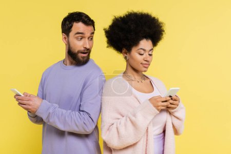 curious man with open mouth looking at african american woman messaging on smartphone isolated on yellow