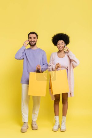 full length of happy interracial couple holding shopping bags and talking on smartphones on yellow background